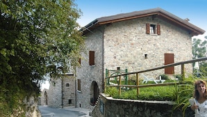 Ein Steinhaus mit geneigtem Dach und einem kleinen Balkon, eine gepflasterte Straße führt dorthin.