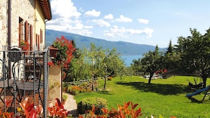 Ein Steinhaus mit Balkon, einem grünen Garten, einer Schaukel und Blick auf See und Berge.