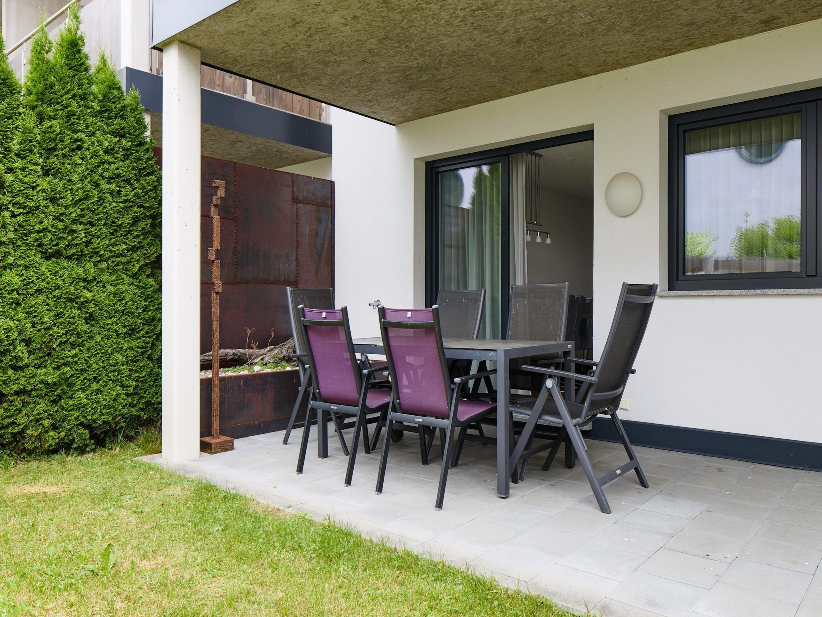 A patio with a table and chairs, a large green hedge, and a metal gate.