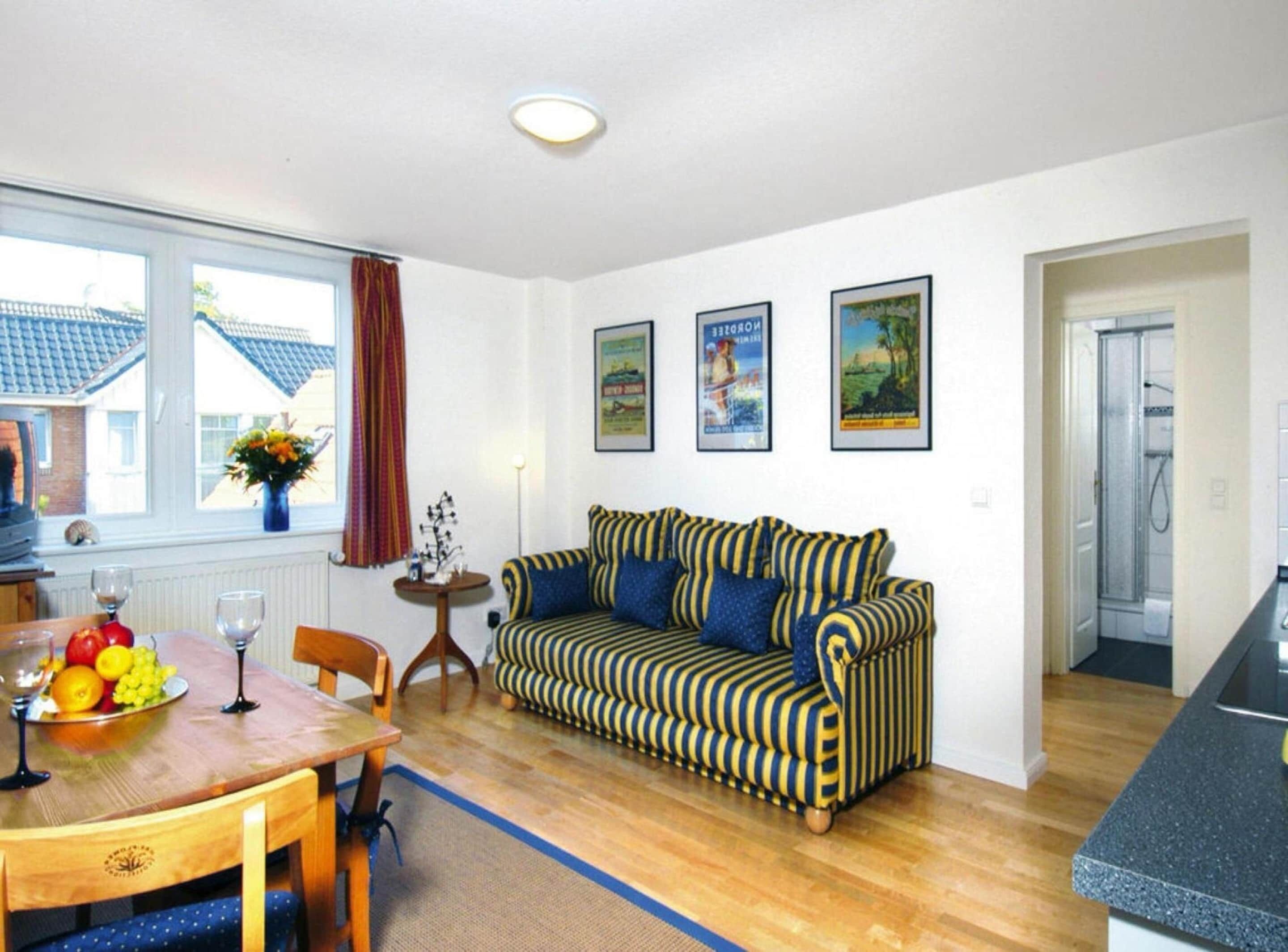 A cozy living room with a striped sofa, a dining table with fruit and wine glasses, and a kitchen area visible through an open door.