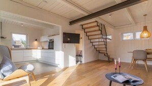 Un espai ample d'estar amb una escala de fusta, una zona de menjador amb taula i cadires, i una cuina amb armaris blancs i un televisor.