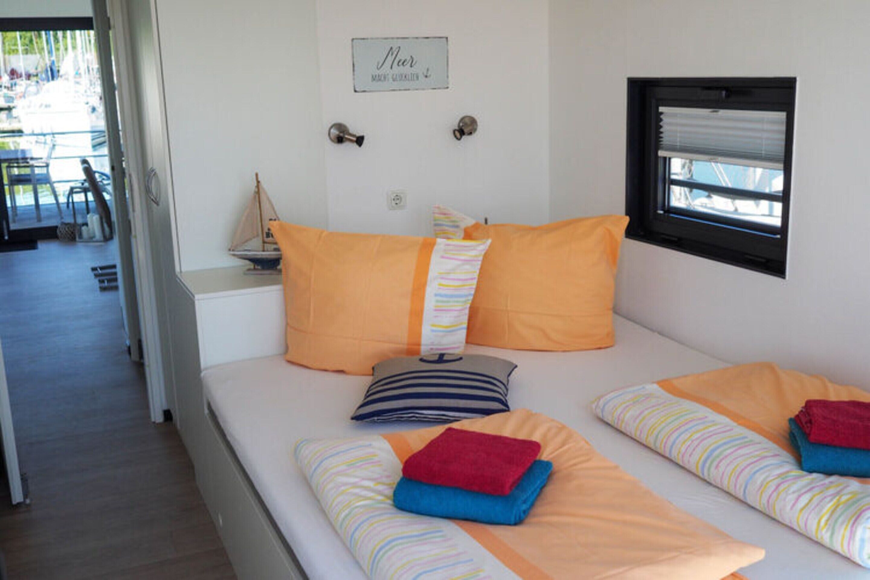 A bedroom with two beds, each with striped bedding and orange pillows. There is a small window with blinds, a door with a sign above it, and a chair visible in the background.