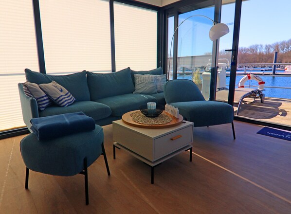 A modern living room with a sofa, two armchairs, a coffee table, and a large window offering a view of a body of water. - Hausboot in Boltenhagen (Boltenhagen)