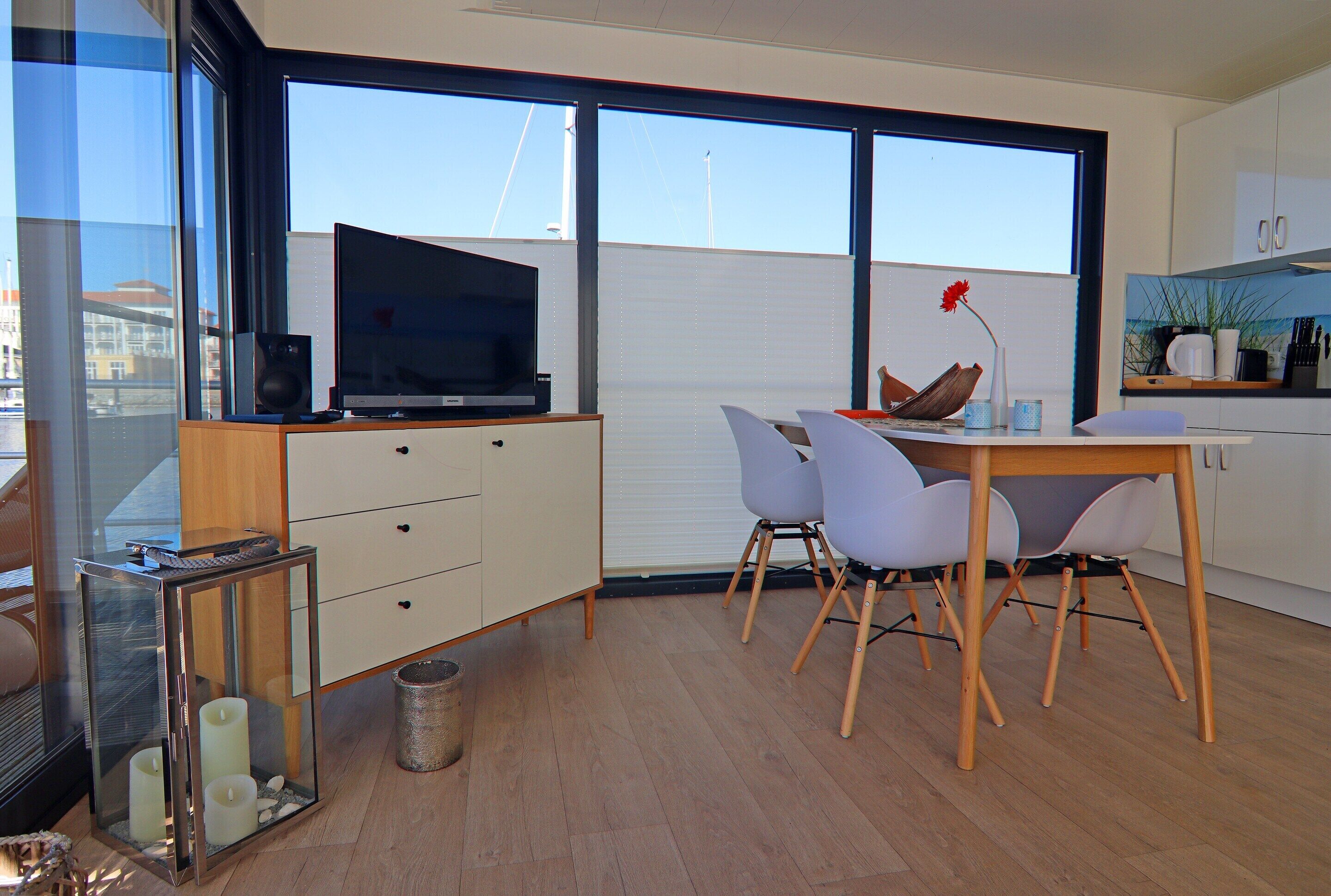 A modern kitchen with white cabinets, a dining area with a wooden table and white chairs, and a large window with a view of the city.