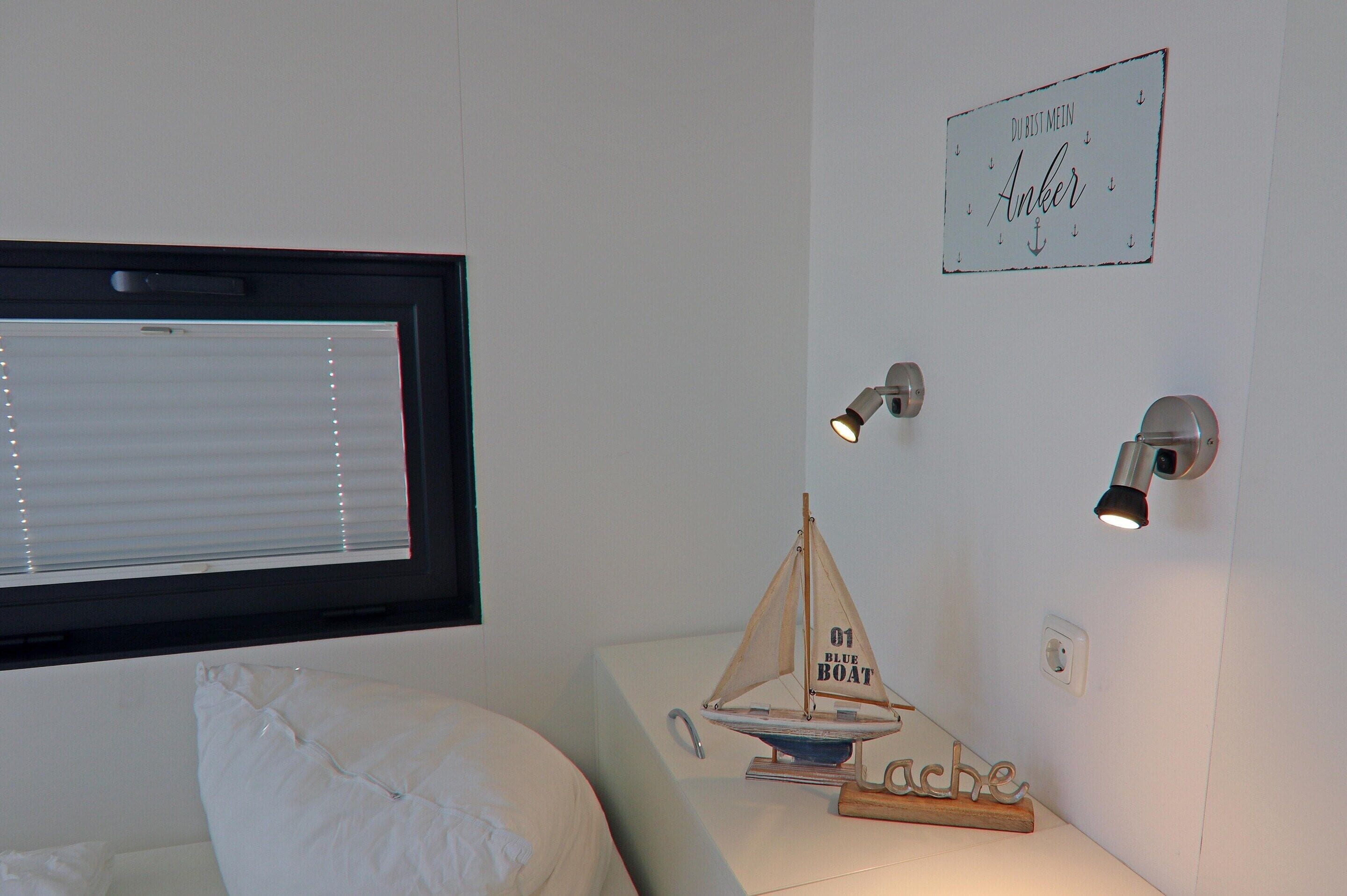 A bedroom with a bed, a window with blinds, a wall-mounted light, a small sailboat decoration, and a sign with text.