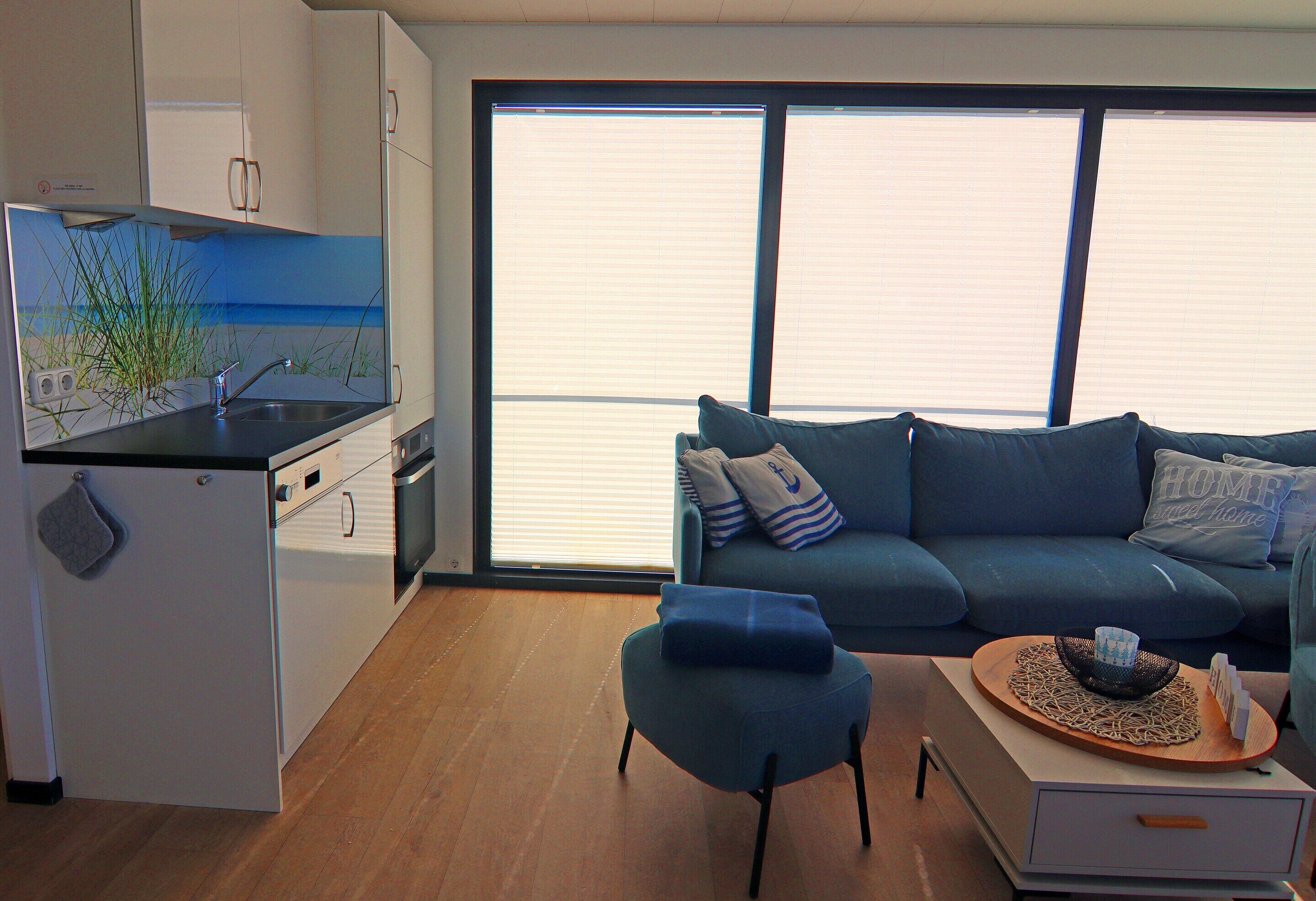 Un espacio habitable moderno con cocina, un sofá azul y un amplio ventanal con persianas.