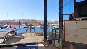 Une pièce avec vue sur un port de plaisance, une terrasse en bois, un bureau, une chaise, une petite table avec des bougies et un tapis bleu orné de motifs.