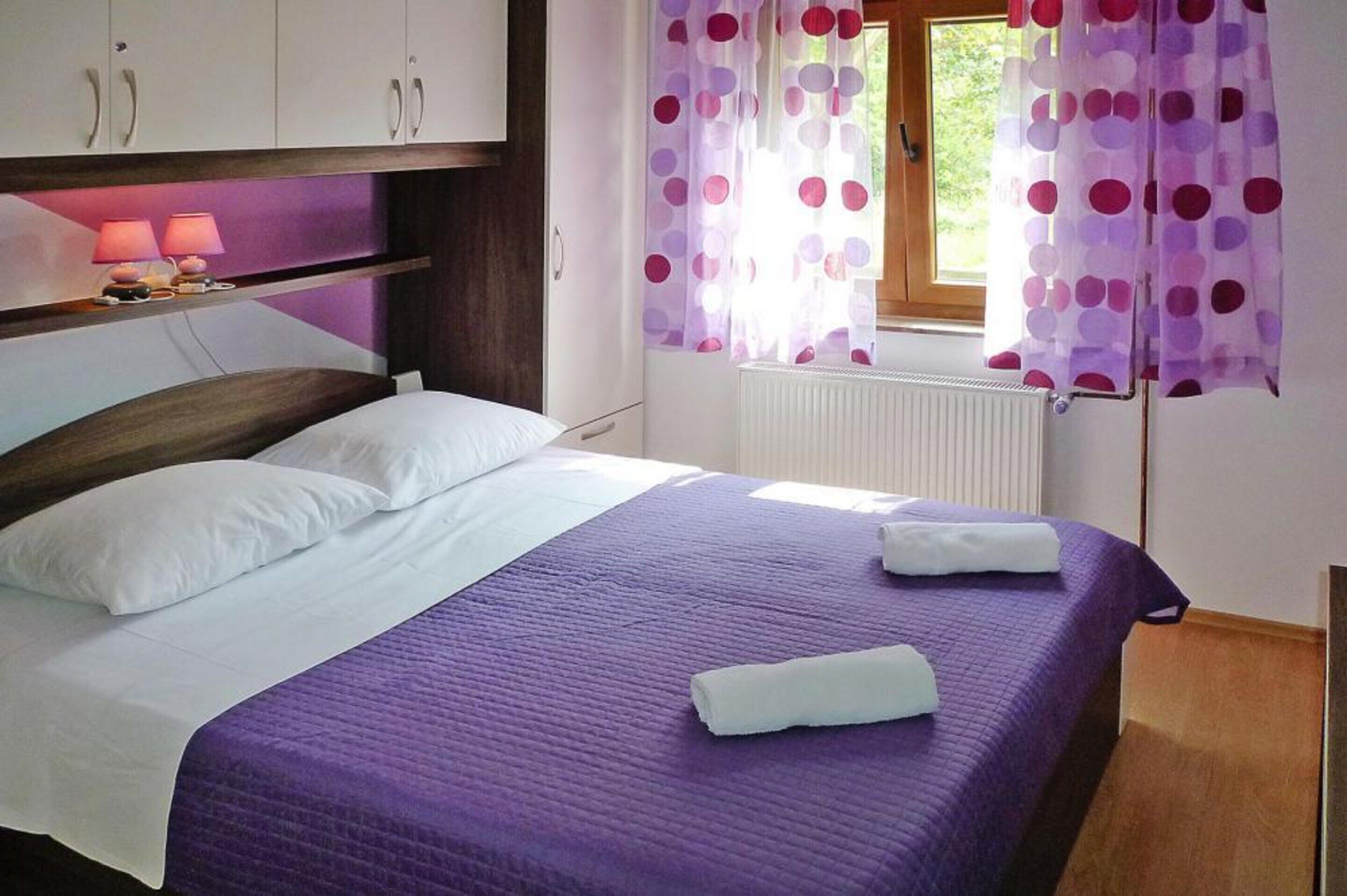 A neatly made bed with a purple bedspread, white pillows, and two folded white towels. There is a window with purple polka dot curtains, a bedside lamp, and a radiator.