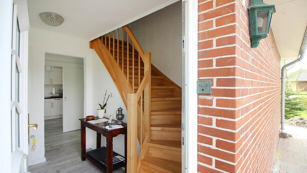 A staircase leading to a second floor, a wooden table with a vase and papers, and a brick wall with a green lantern. - Cottage, Oldenswort (Oldenswort)