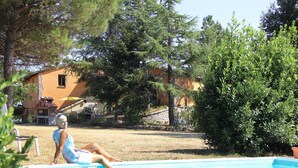 Una persona relajándose junto a una piscina, con una casa y árboles al fondo.