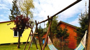 Un hamac tendu entre deux poteaux en bois, un bâtiment jaune et un bâtiment rouge, le tout dans une cour verdoyante.