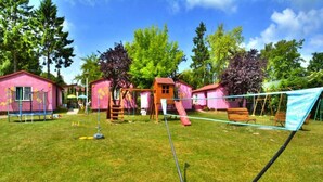 Une cour verdoyante avec un trampoline, une balançoire et un toboggan. On aperçoit deux bâtiments roses au fond.