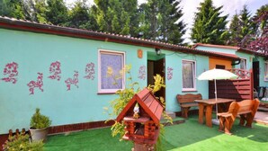 Une maison à l’extérieur verdoyant, dotée d’une terrasse en bois et d’un petit jardin agrémenté de gazon synthétique et de plantes en pot.