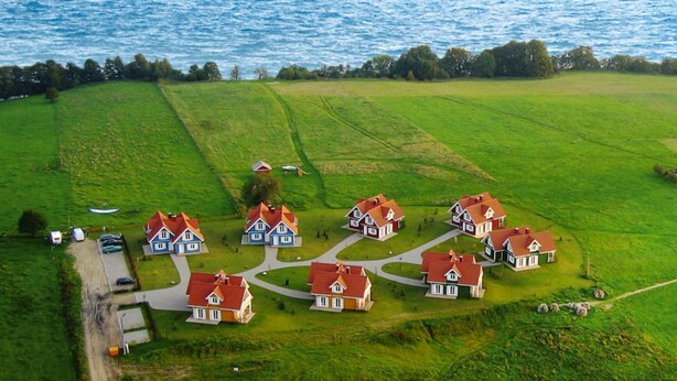 An aerial view of a coastal village with houses arranged in a circular pattern around a central green space, with a road leading to the sea. - Cottage Directly by the Lake in Trygor (Wegorzewo)