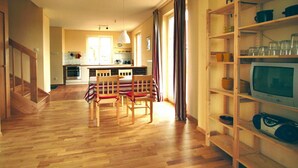 A spacious living area with a wooden floor, a dining table with chairs, a kitchen in the background, and a TV on a shelf. - Cottage Directly by the Lake in Trygor (Wegorzewo)