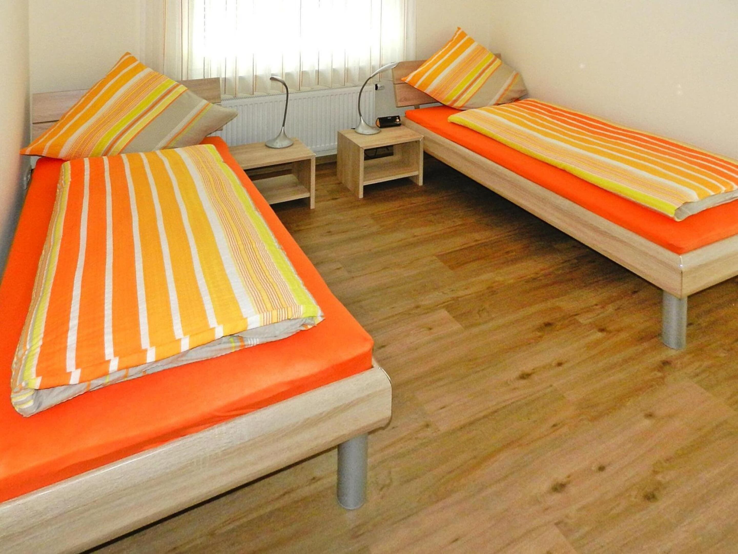 Ein Zimmer mit zwei Einzelbetten, jeweils mit einer orangefarbenen Matratze und gestreiften Kissen. Dazwischen befindet sich ein kleiner Nachttisch mit einer Lampe.