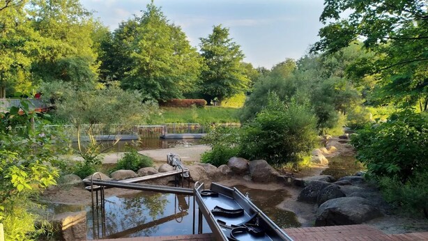 A tranquil outdoor pool area with a metal slide and a small pond, surrounded by lush greenery and trees. - Reihenhaus in Lubben an der Spree (Lübben (Spreewald))