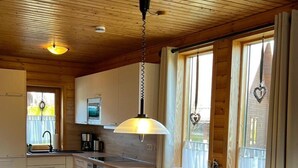 Ein gemütliches Blockhausszimmer mit Holzbalkendecke, einem runden Esstisch mit vier Stühlen, einer Küchenzeile mit Herd und Schränken sowie großen Fenstern mit Vorhängen.