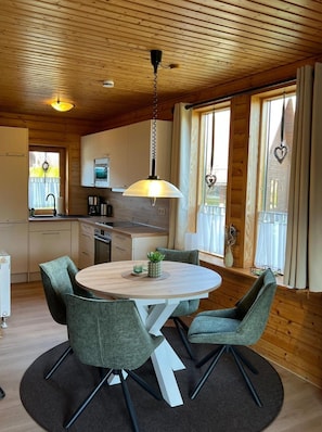 Ein gemütliches Blockhausszimmer mit Holzbalkendecke, einem runden Esstisch mit vier Stühlen, einer Küchenzeile mit Herd und Schränken sowie großen Fenstern mit Vorhängen.