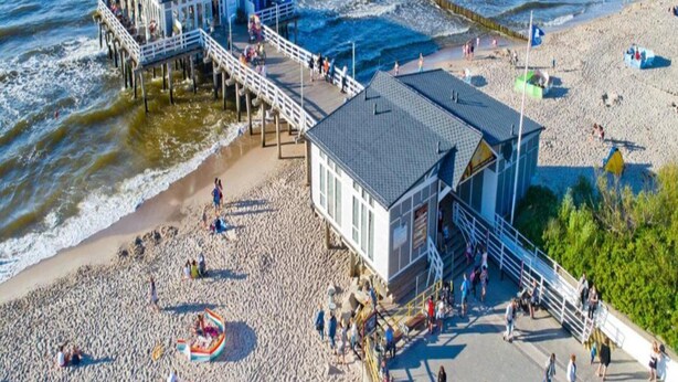 Eine Steg, der ins Meer führt, mit einem Strandabschnitt und einem Rettungsschwimmerstand.