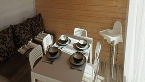 Eine Holzhütte mit Essbereich, ausgestattet mit Tisch, Schüsseln und Tellern, weißen Plastikstühlen und einer Couch mit gemusterten Kissen.