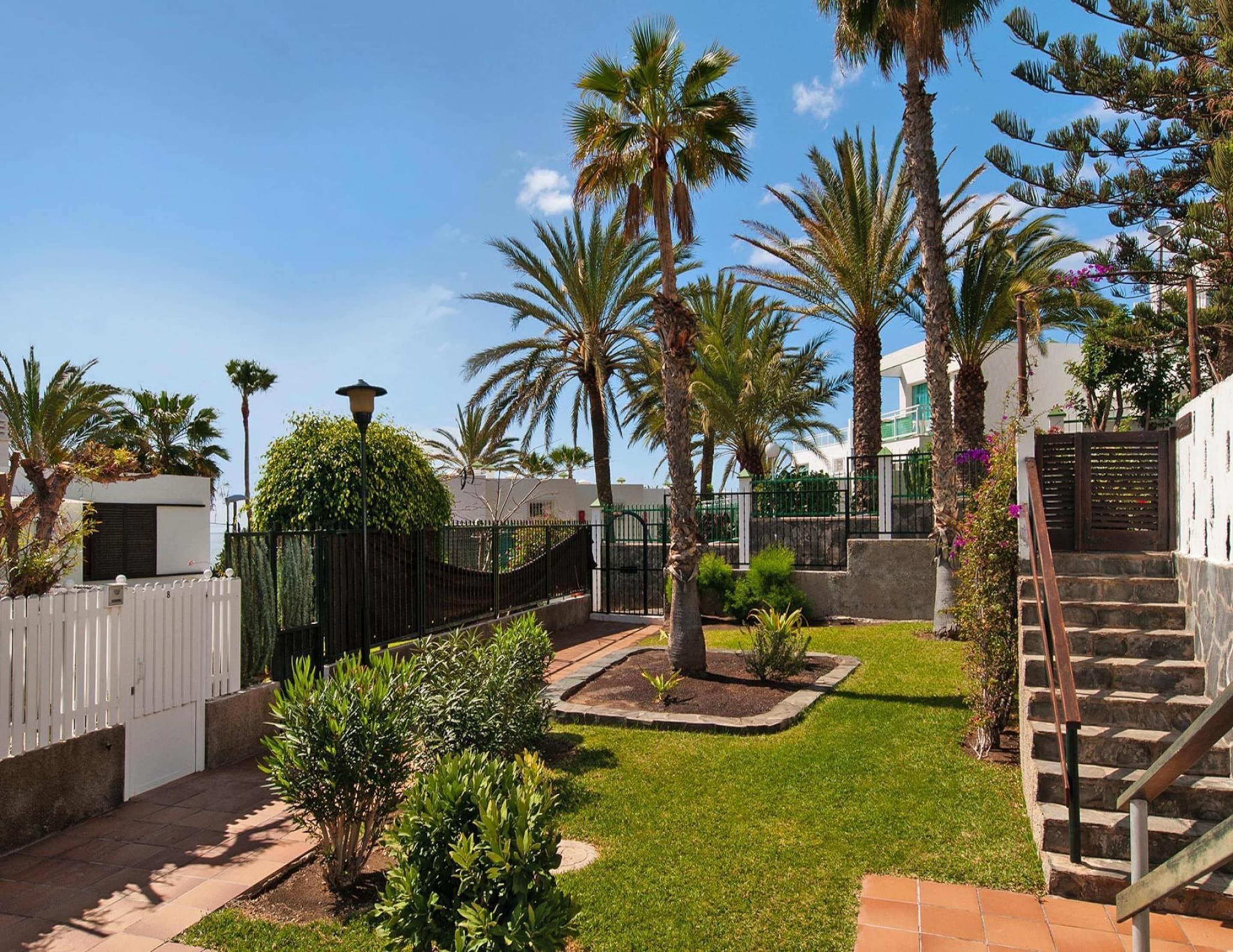 Un jardín bien cuidado con palmeras, una valla blanca y una escalera de piedra que lleva a un edificio.