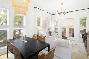 A bright, open-plan living area with a dining table, wicker chairs, a white sofa, and a chandelier.
