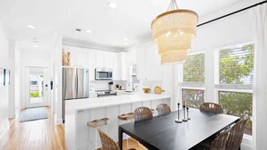 Una cocina moderna con muebles blancos, electrodomésticos de acero inoxidable y un comedor con una mesa oscura y sillas de mimbre.