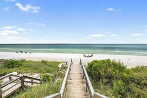 Un paseo marítimo de madera que conduce a una playa con aguas transparentes y algunas personas a lo lejos.