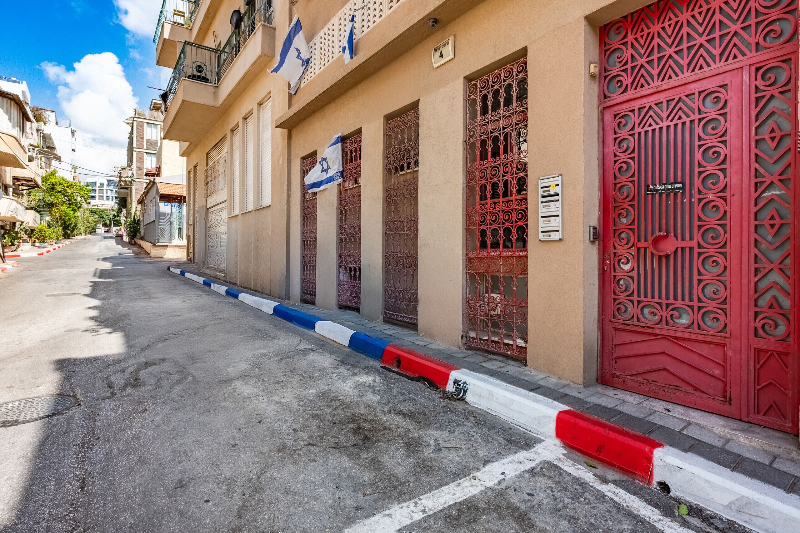 Une vue de rue avec une rangée de bâtiments, dont certains sont dotés de balcons et de drapeaux, et une porte rouge ornée de motifs complexes.