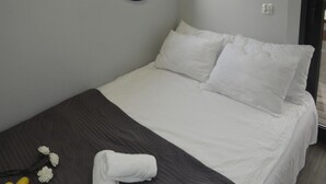 Ein ordentlich bezogenes Bett mit weißen Leinen, einer dunkelbraunen Tagesdecke und einem weißen Kopfkissen. Zwei weiße Blumen und ein aufgerolltes weißes Handtuch liegen auf dem Bett.