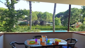 A balcony with a table set for two, surrounded by chairs, overlooking a swimming pool and a well-manicured garden. - Villas Nautilus in Bibione With Pool (San Michele al Tagliamento)
