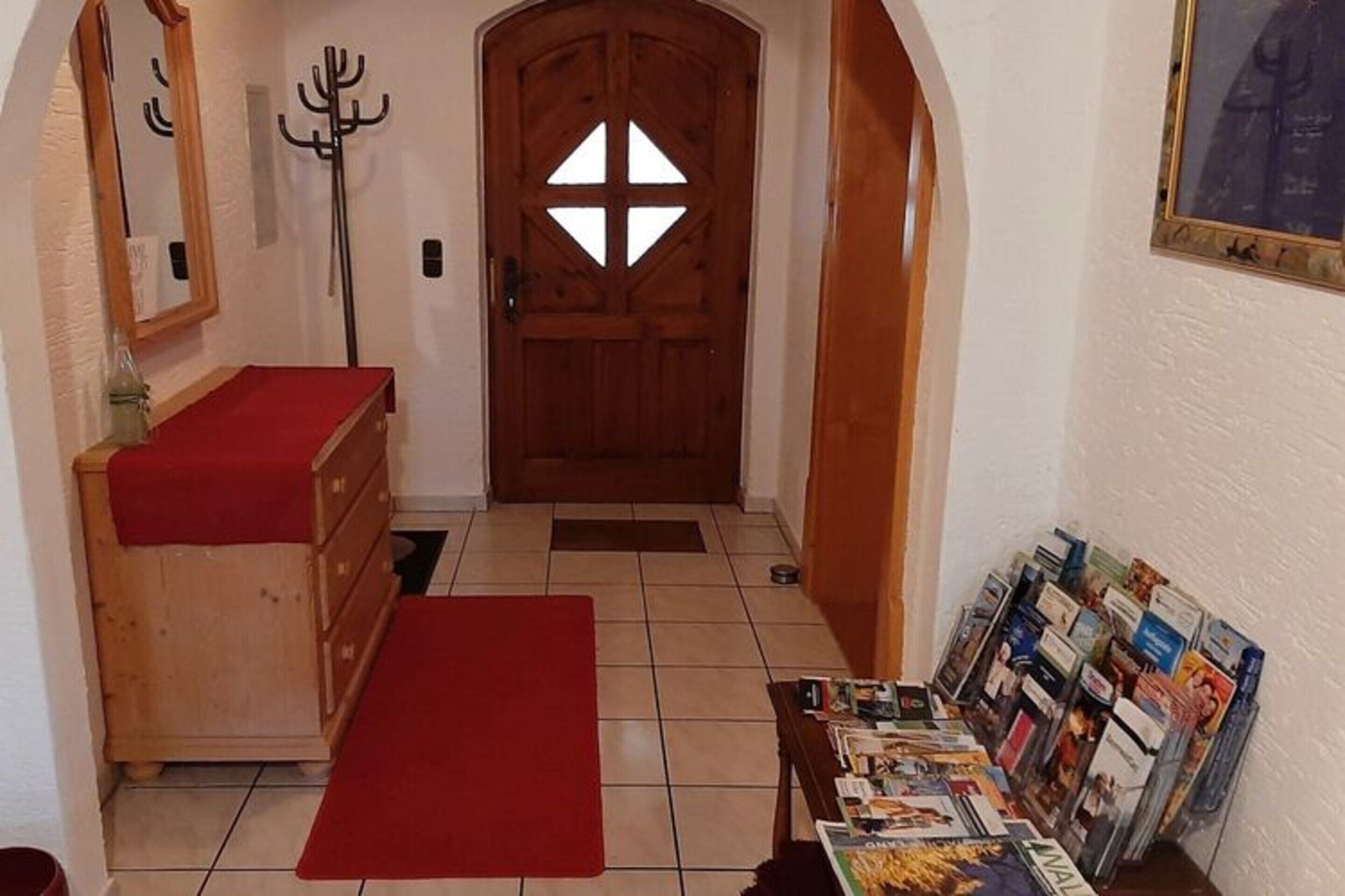 Un pasillo con una puerta de madera, un perchero, un banco de madera con un cojín rojo y una mesa con revistas.