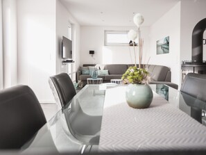 Een moderne woonkamer met een glazen eettafel, een grijze bank en een aan de muur bevestigde televisie.