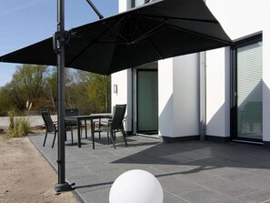 Een terras buiten met een tafel, stoelen, een zwarte parasol en een witte, bolvormige vorm.