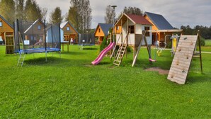 Ein Spielplatz mit Rutsche, Schaukel und Kletterbereich in einer grünen Wiese, im Hintergrund hölzerne Häuser.