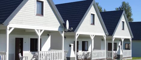 Une rangée de cabanes blanches aux toits sombres, chacune dotée d’un porche avec des balustrades blanches et de sièges d’extérieur.