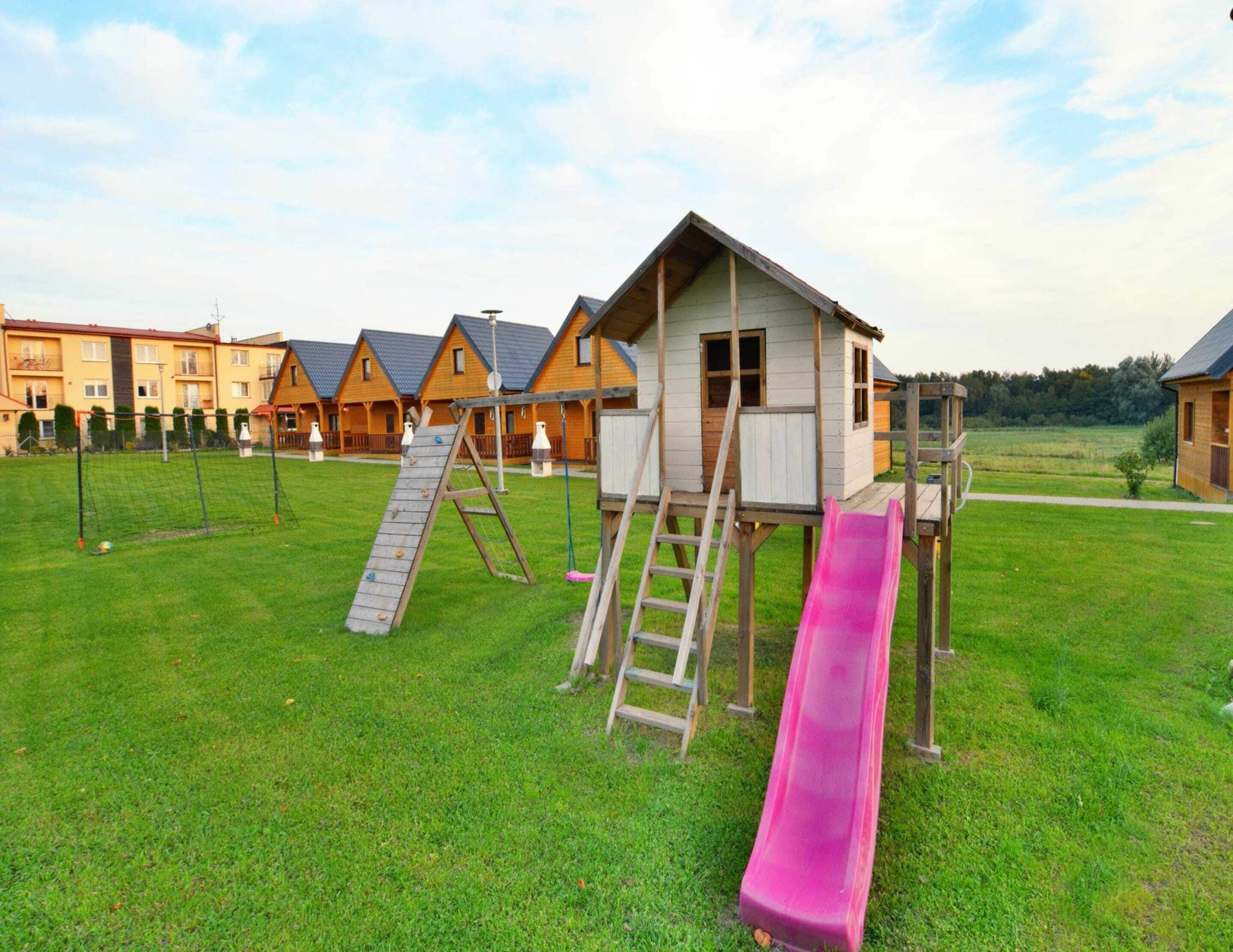 A playground with a slide and climbing structure in a grassy area with wooden buildings in the background.