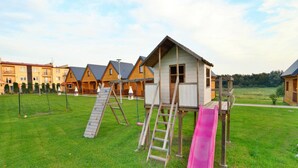 Ein Spielplatz mit Rutsche und Klettergerüst, umgeben von Rasen, mit Holzbauten im Hintergrund.