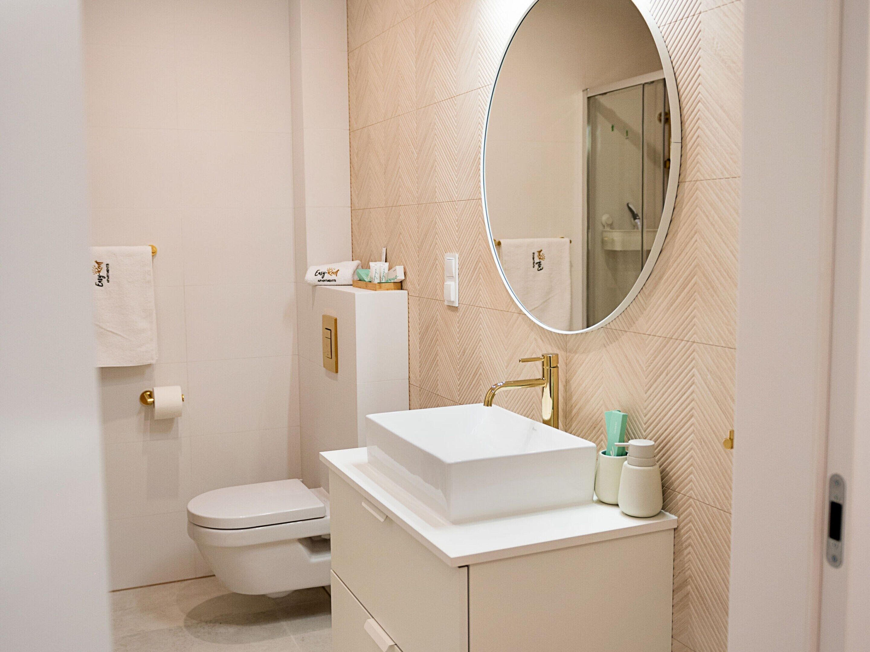 A modern bathroom with a white sink, gold faucet, and a mirror above it. There is a toilet to the left and a towel rack above it.