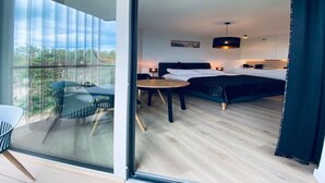 Una habitación de hotel moderna con un gran ventanal corredero que da a un balcón, una cama con cabecera negra, una pequeña mesa y una silla.