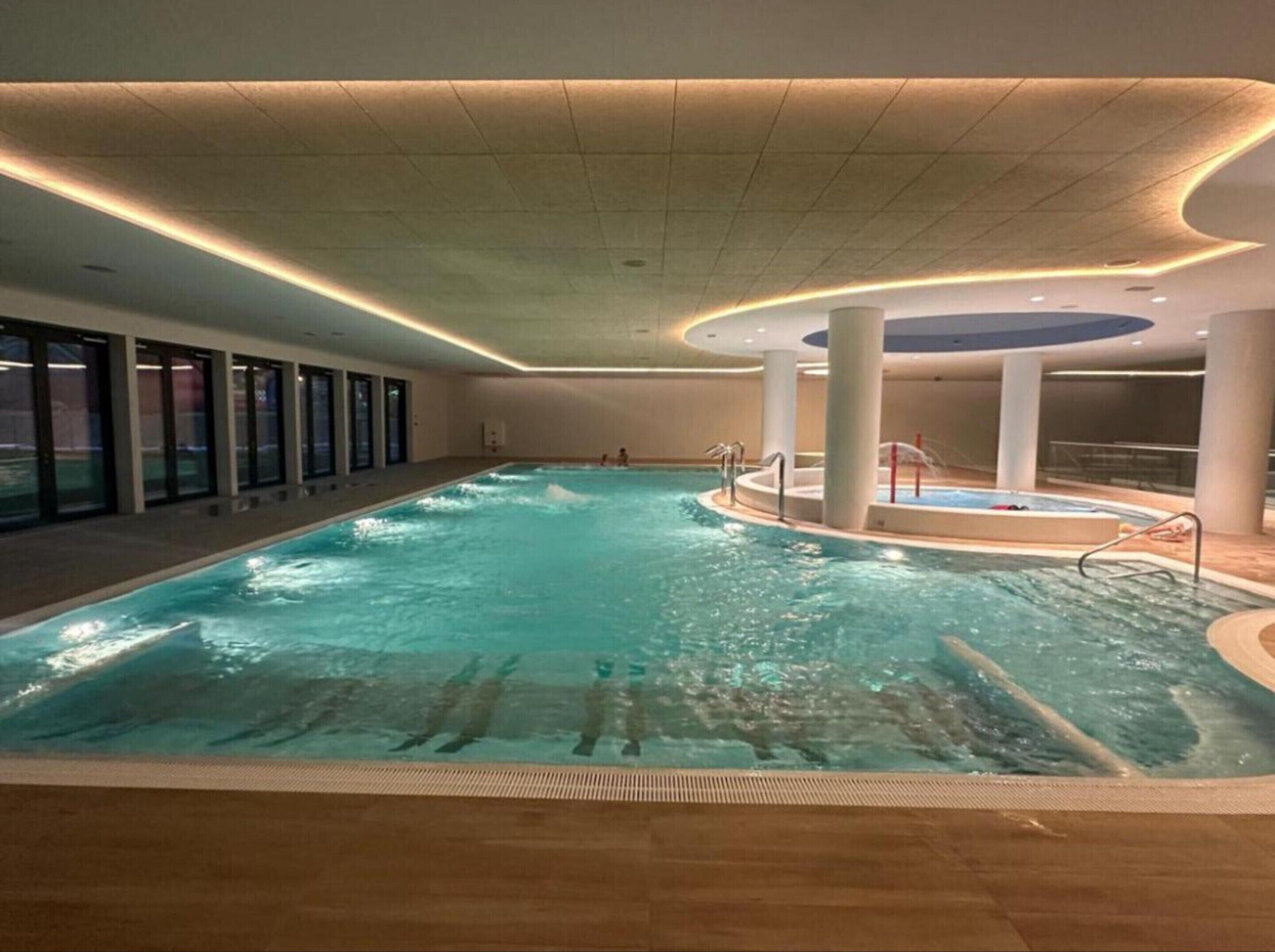 Una piscina interior con agua cristalina, rodeada por un muelle de madera y paredes de vidrio.