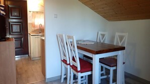A dining area with a wooden table and white chairs, a kitchen in the background, and a sloped ceiling. - Holiday Home in Kolczewo With Lake Access (gmina Wolin)