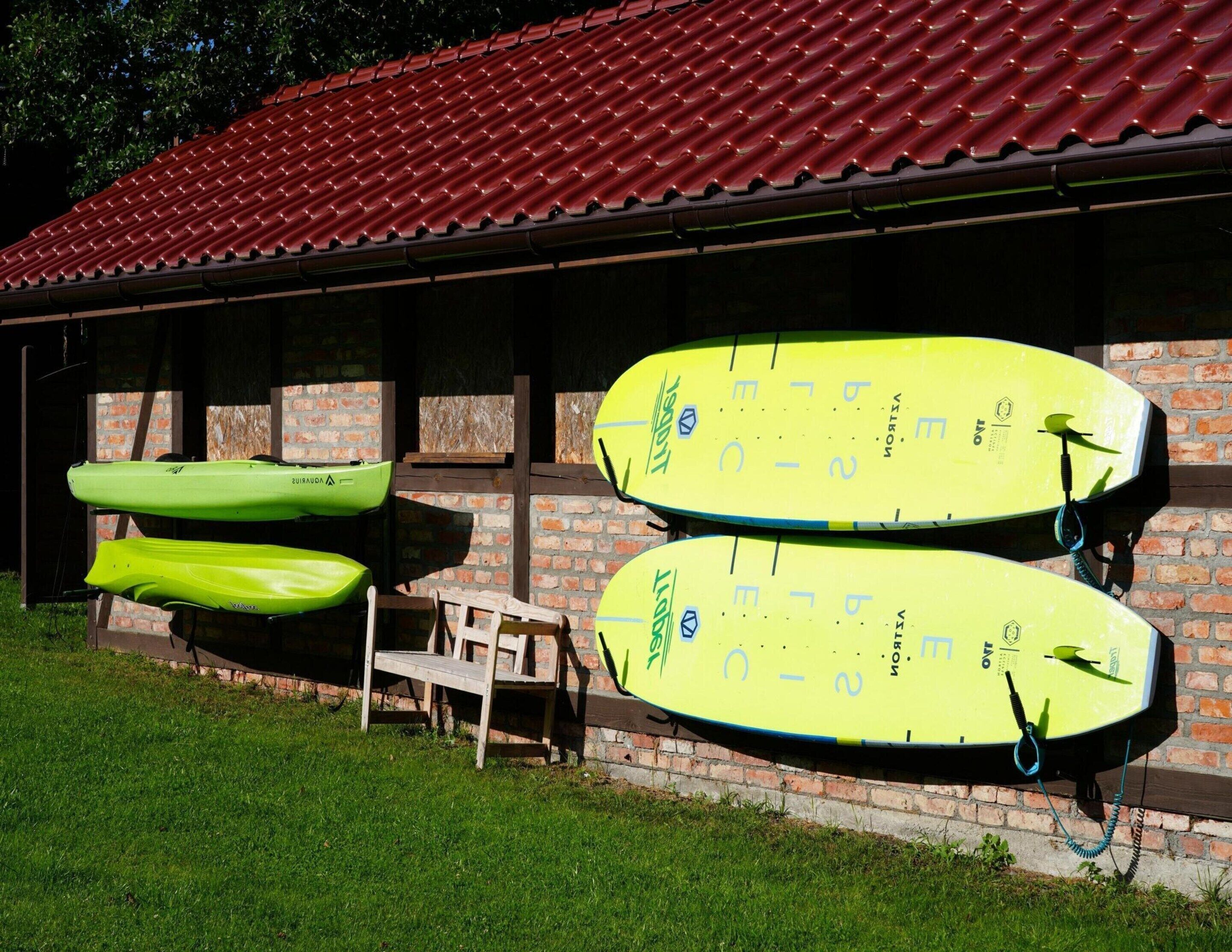 Two kayaks and a paddleboard leaning against a brick wall with a red-tiled roof.