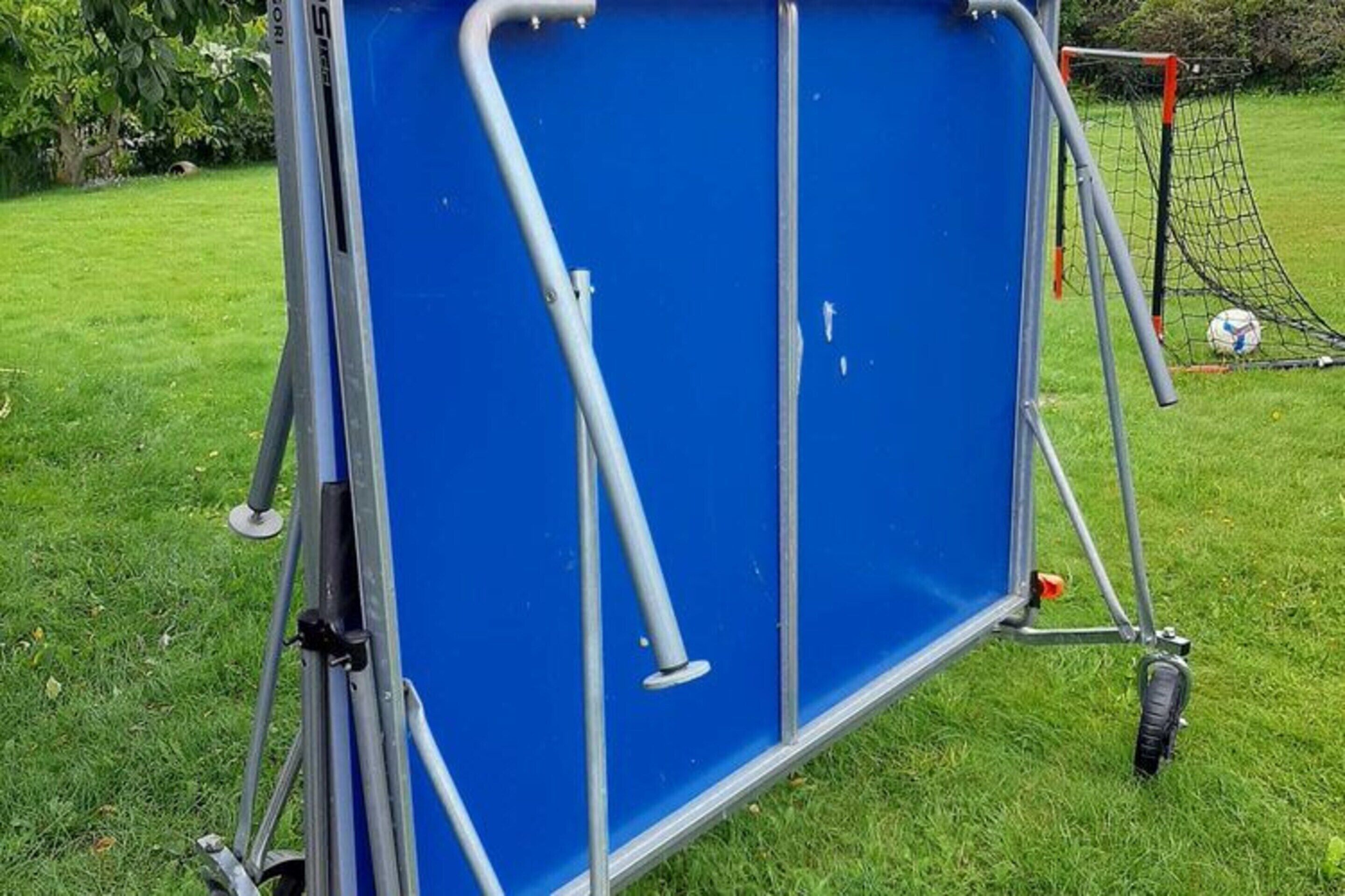 Une structure métallique bleue, dotée de poteaux et de roues, vraisemblablement un but de fortune ou un espace de rangement mobile, située dans un espace extérieur recouvert d’herbe.