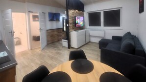Un salon moderne avec une table à manger en bois, des canapés noirs, un téléviseur et un mur d’accent en pierre.