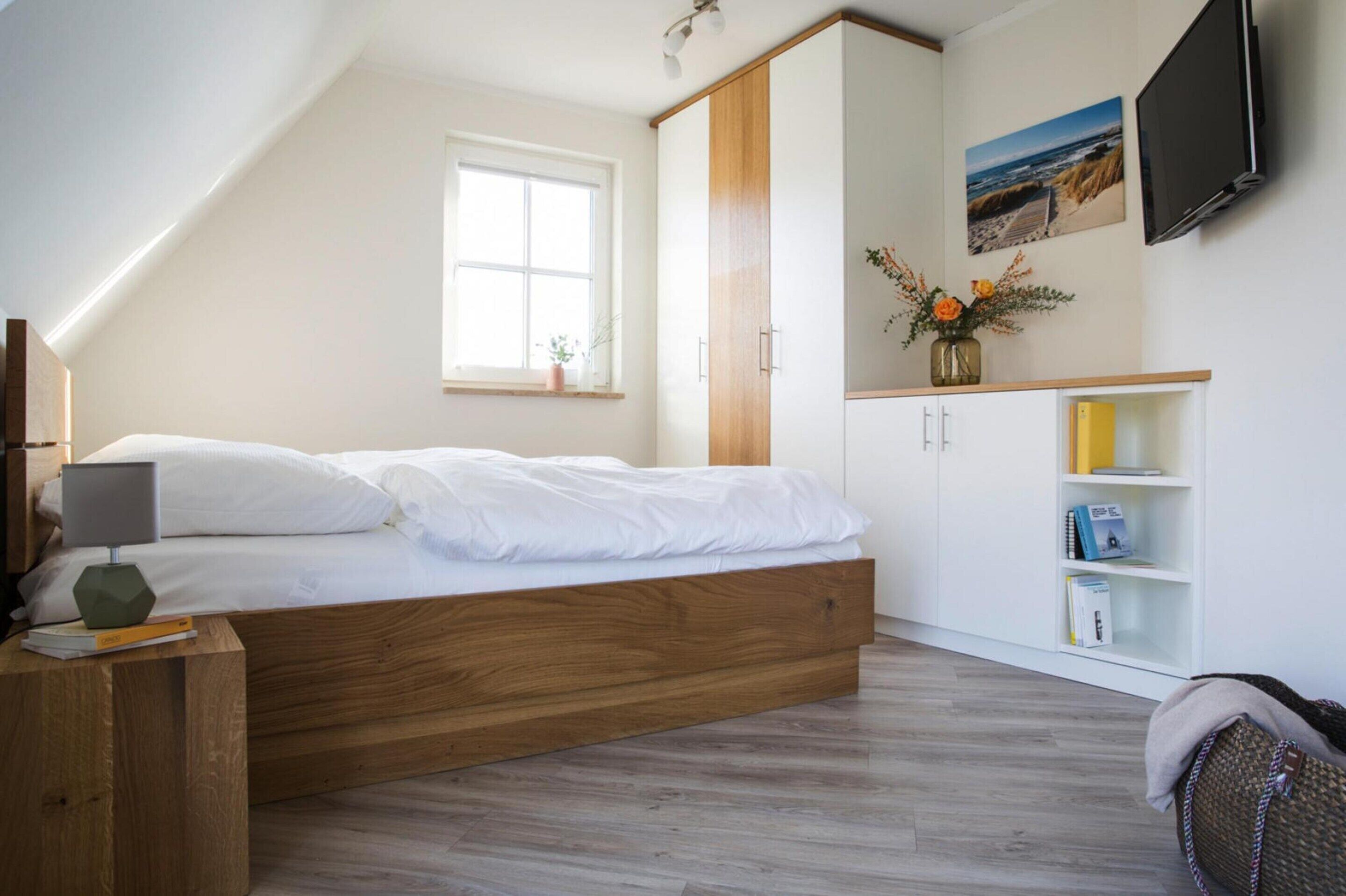 A bedroom with a wooden bed, a white wardrobe, a TV mounted on the wall, and a window with a view of the outdoors.