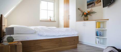 A bedroom with a wooden bed, a white wardrobe, a TV mounted on the wall, and a window with a view of the outdoors.