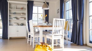 A bright dining area with white furniture, a table with chairs, a hanging light, and a bookshelf.
