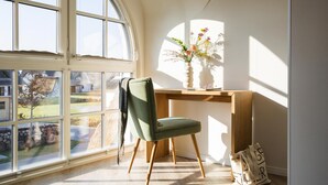 A room with a large window, a green chair, a wooden desk, and a vase with flowers.